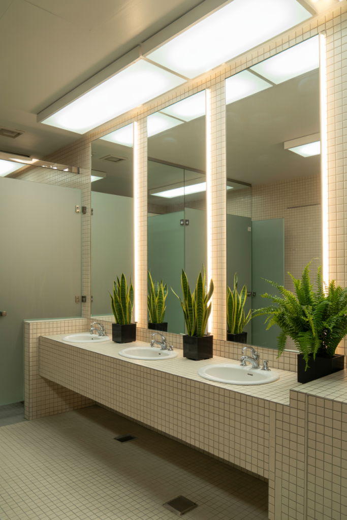 A modern public restroom with beige tile, three sinks, large mirrors with glowing lights, and potted green plants placed between the sinks. Stalls with frosted doors are visible in the background.