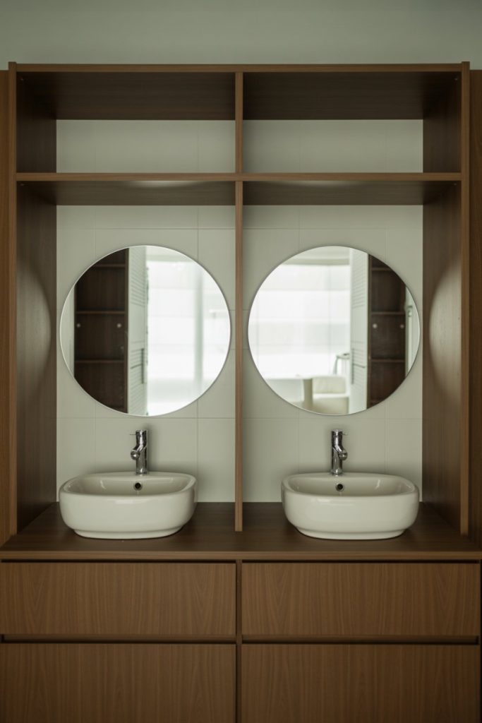 A modern bathroom with two round mirrors above two white sinks set in a wooden vanity, featuring open shelves and drawers beneath the sinks. The background shows a tiled wall reflected in the mirrors.