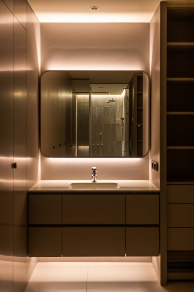 Modern bathroom with warm recessed lighting, a large rectangular mirror above a sink with a single faucet, minimalistic cabinetry, and open shelving. The reflection shows a glass shower in the background.