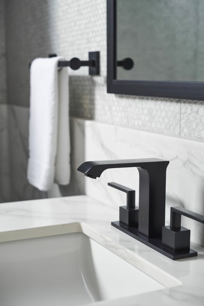 A modern bathroom with a black faucet on a white marble sink, a towel hanging on a black rod, and a large mirror with a black frame on a tiled wall.