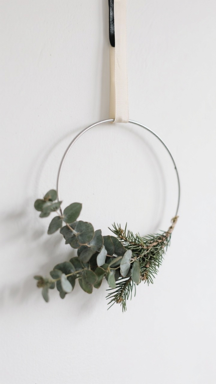 Closeup portrait of a minimal asymmetrical wreath on a white wall: slim metal hoop with eucalyptus and cedar clustered on one-third of the circle, secured with floral wire concealed by a neat ribbon; hung from black leather or cream ribbon; shallow depth of field, clean Scandinavian background.